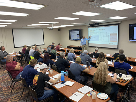 Man teaching about asphalt on a large screen in the front of a room with 30 people at tables.